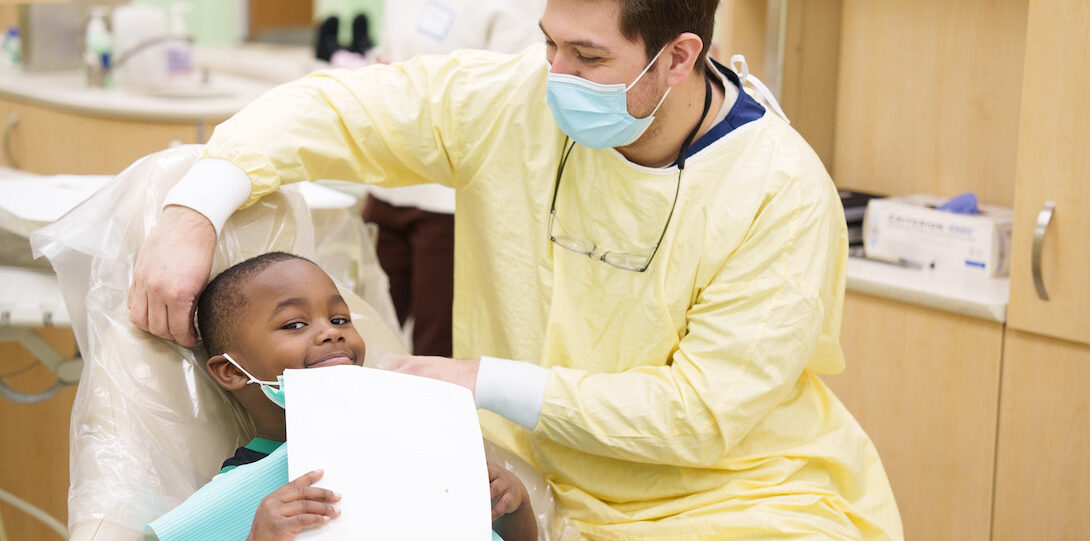 A dental student in yellow scrubs giving a young student a teeth cleaning