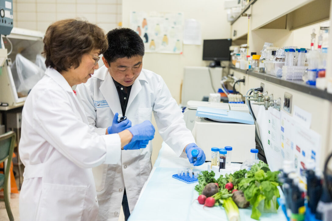 Dr. Wu working in lab and researcher both wearing white coats, in front of a pile of vegetables