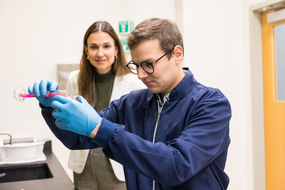 Dr. Russo in white lab coat standing behind a student dressed in blue, who is pouring a pink liquid