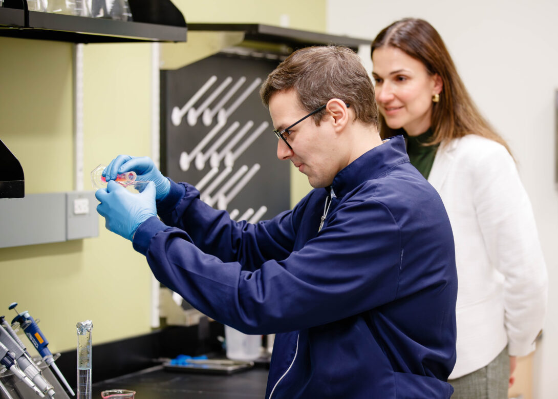 man wearing navy blue pouring solution into beaker, woman in lab coat stands behind him