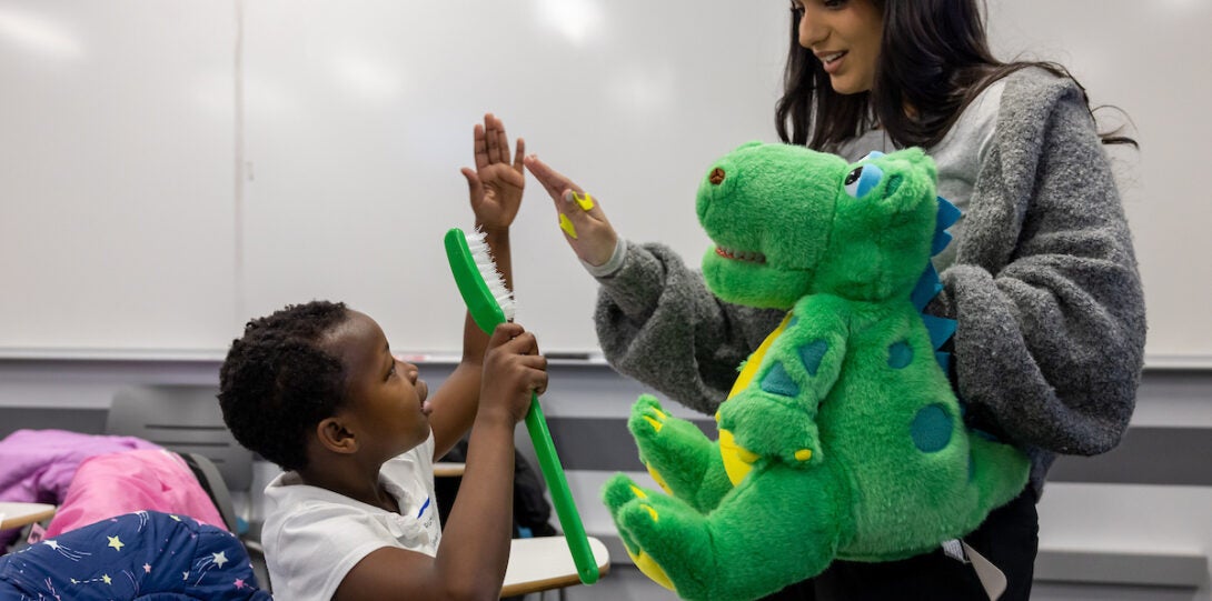 School child with oversized toothbrush hi-fives pre-dental volunteer with green dinosaur puppet